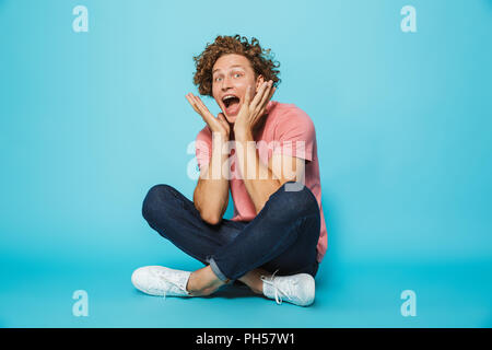 Portrait d'un jeune homme aux cheveux bouclés joyeux criant assis les jambes croisées sur fond bleu Banque D'Images