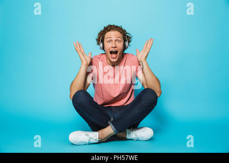 Portrait d'un jeune homme aux cheveux bouclés excité dans les écouteurs assis les jambes croisées sur fond bleu Banque D'Images