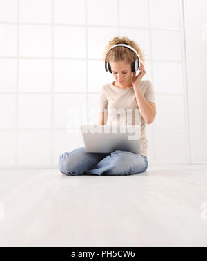 Jeune femme avec l'ordinateur et les écouteurs, regardez le moniteur portable, assis sur le plancher isolé sur fond blanc fenêtre chambre Banque D'Images