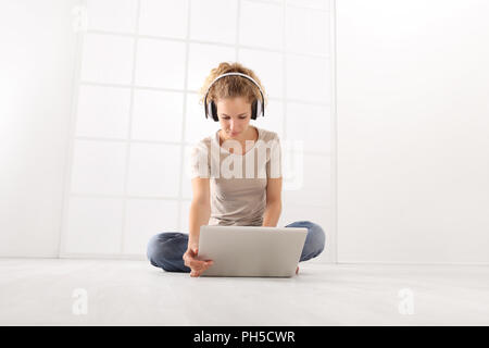 Jeune femme avec l'ordinateur et les écouteurs, regardez le moniteur portable, assis sur le plancher isolé sur fond blanc fenêtre chambre Banque D'Images