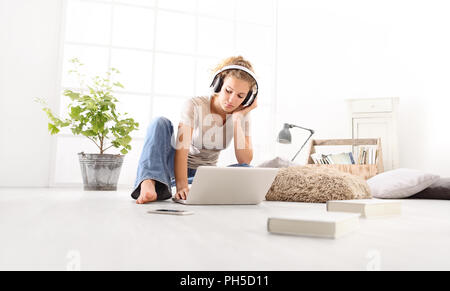 Jeune femme avec ordinateur, casque, smartphone et books, assis sur le plancher dans la salle de séjour on white large fenêtre à l'arrière-plan Banque D'Images
