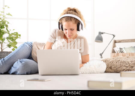 Jeune femme avec ordinateur, casque, smartphone et livres, gisant sur le sol dans la salle de séjour on white large fenêtre à l'arrière-plan Banque D'Images