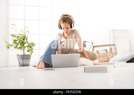 Jeune femme avec ordinateur, casque, smartphone et books, assis sur le plancher dans la salle de séjour on white large fenêtre à l'arrière-plan Banque D'Images