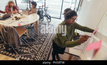 Jeune femme écrivant sur un tableau blanc au démarrage de l'office. Les modalités d'un nouveau plan de projet sur un tableau blanc avec des collègues travaillant sur un ordinateur portable à l'arrière. Banque D'Images