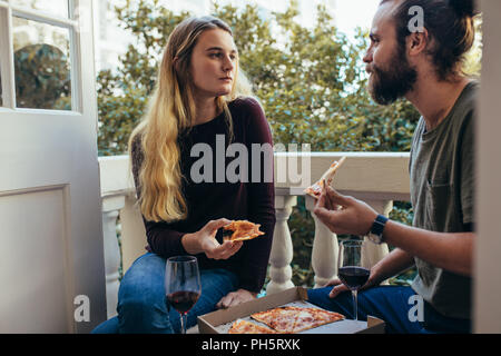 Couple partageant une pizza assis dans le balcon de leur appartement. L'homme et la femme assis ensemble et de manger une pizza avec du vin. Banque D'Images
