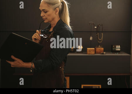 Portrait of mature femme créateur de bijoux à la recherche à la conception d'un livre. Femme joaillier à tablier avec livre design par l'atelier. Banque D'Images