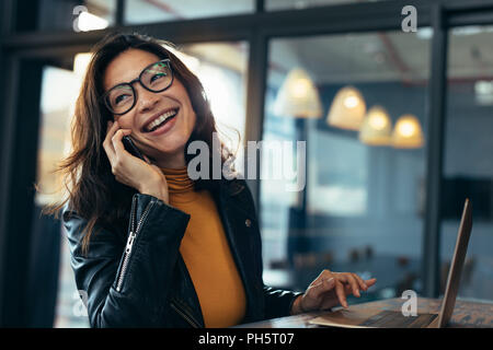 Cheerful asian female talking on mobile phone, assis sur un bureau avec ordinateur portable. Business Woman in casuals effectuer un appel téléphonique et de rire. Banque D'Images