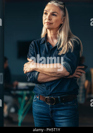 Portrait de senior business woman standing in office avec les bras croisés à l'écart et la pensée. Thoughtful female business owner in office. Banque D'Images