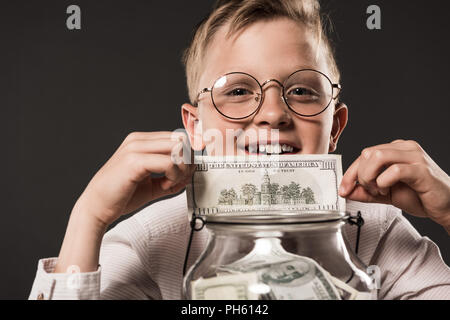 Portrait of smiling little boy lunettes en montrant plus de billets en dollars pot plein de cash money isolé sur fond gris Banque D'Images