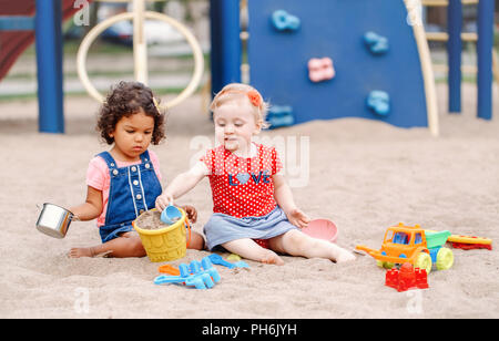 Partager avec un ami. Deux blancs et hispaniques mignon Amérique Bébés Enfants assis sur sable à jouer avec des jouets colorés en plastique. Peu de filles ayant Banque D'Images