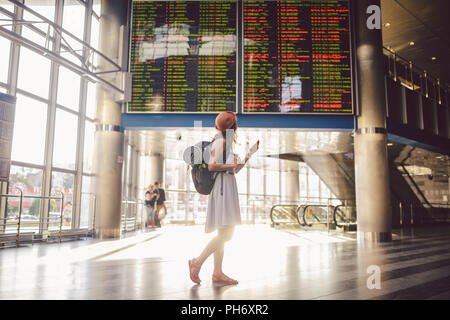 Voyages à thème et tranosport. Beau young caucasian woman in dress et son sac à dos se tenant debout à l'intérieur gare ou terminal à un planning à tenir un téléphone rouge, utilise la technologie de la communication Banque D'Images