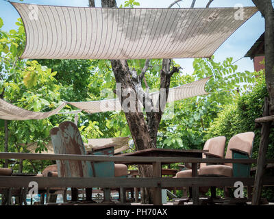 Vintage fauteuils avec table en bois sur la terrasse en bois avec toit en tissu vert sur fond de ciel bleu et des arbres. Mix and Match unique de style meubles Banque D'Images