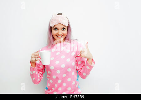 Bonjour. Souriante jeune femme montre pouce vers le haut. Girl in pink pyjamas et masque de sommeil sur fond blanc Banque D'Images