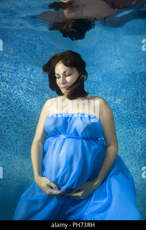 Pregnant woman in a blue dress posing sous l'eau dans la piscine Banque D'Images