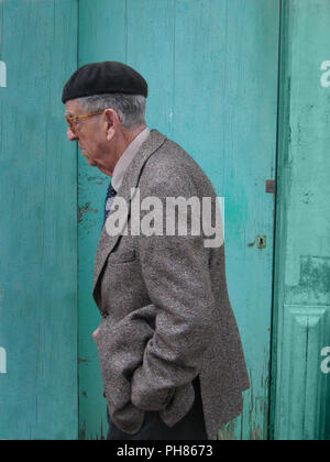 Portrait de profil d'un vieil homme portant un béret espagnol et une veste et une cravate de sport grises élégantes. Il marche au-delà d'une porte en bois turquoise dans une rue à Sant Lluis, Minorque, Espagne. Le vieil homme semble un peu inquiet et préoccupé Banque D'Images