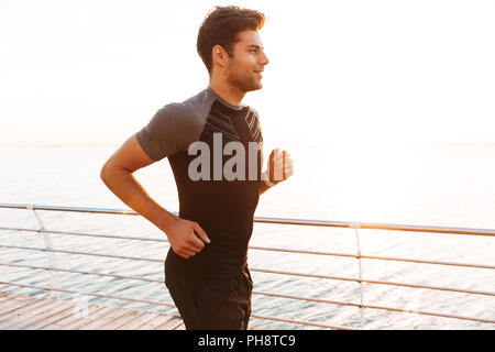 Photo de guy sportive européenne 20s en survêtement jogging le long du quai ou promenade de mer au lever du soleil Banque D'Images