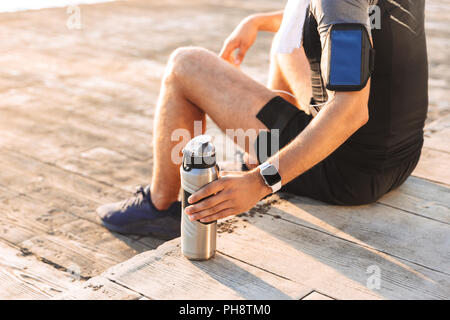 Photo recadrée de runner 20s l'homme en survêtement avec le support de smartphone assis sur jetée en bois au bord de mer et l'eau potable à partir de la Tasse thermos Banque D'Images