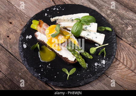 Pain grillé avec des bâtons d'asperges vertes grillées enrobé de bacon, oeuf poché , pousses et de fromage bleu sur noir sur ardoise rustique en bois ancien Banque D'Images