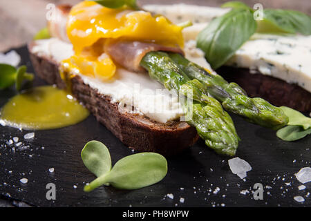 Pain grillé avec des bâtons d'asperges vertes grillées enrobé de bacon, oeuf poché , pousses et de fromage bleu sur noir ardoise pierre close up. Banque D'Images