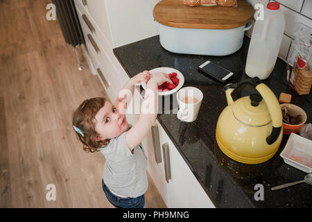 Portrait d'une petite fille dans un standning et possèdent jusqu'au comptoir de la cuisine pour prendre une bouteille de framboises. La petite fille est à la recherche Banque D'Images