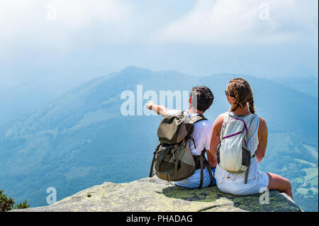 Les jeunes touristes couple avec backpaks assis sur un bord d'une montagne bénéficiant d'une vue panoramique. Banque D'Images