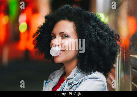 Jolie jeune femme aux cheveux très bouclés se gonfle une bulle de chewing-gum ball blanc la nuit. Trendy girl s'amuser, profiter de la vie. Belle il Banque D'Images