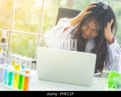 Young Asian female scientist assis sur une chaise tenant sa tête avec la main et visage sérieux parce que la pression dans son travail avec l'équipement, microscope, Banque D'Images