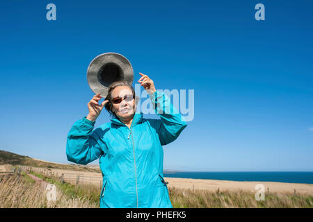 Attrayant, Mature Asian lady wearing blue anorak, une toile chapeau et lunettes de soleil, son chapeau s'envole pas dans le vent fort. Banque D'Images