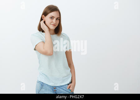 Peut-on parler pour le moment. Mignon et timide jeune brunette attrayant doux avec coupe courte d'effleurement de cheveux derrière l'oreille et le regard tendre à huis clos, avec légère beau sourire posant sur fond gris Banque D'Images