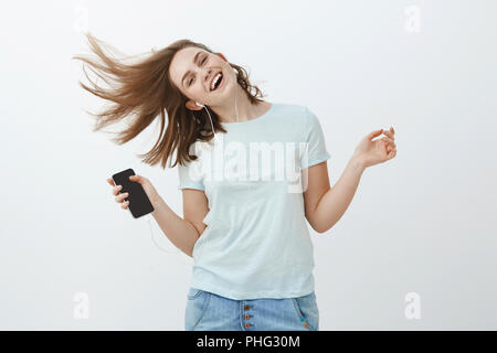 La musique est une grande émotion booster. Portrait de femme joyeuse et charmante emotive agitant joyeusement le saut de joie et sourire à l'écoute de la musique dans des écouteurs holding smartphone posant contre mur gris Banque D'Images