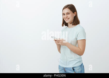 Ne partez jamais sans musique. Portrait de charmant friendly-ravi à la jeune femme en tenue décontractée se préparer à pied à l'université portant des écouteurs et le sourire de côté holding smartphone Banque D'Images