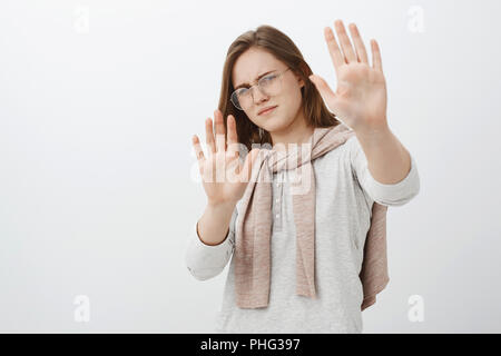 L'étape retour vous dérange pas. Cute femme intense déplu dans les verres avec les cheveux bruns courts massifs en arrière la palms en défense en essayant de protéger les yeux du soleil ou lampe sur mur gris Banque D'Images