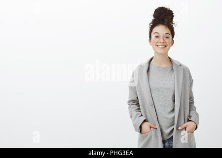 Portrait of happy bon-à la fille aux cheveux bouclés peignées à bun, le port de lunettes et manteau gris, tenant les mains dans les poches et sourire gaiement à l'appareil photo, se sentir plus impressionnant mur gris Banque D'Images