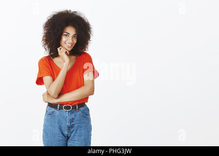 Studio shot of African American girl flirty magnifique avec des cheveux bouclés, souriant avec charmante expression et tenant la main près du cou, être intrigué et intéressé à ce jour avec mignon guy sur mur gris Banque D'Images