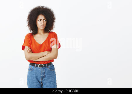 Tourné à l'intérieur attrayant de femmes à l'outcast avec coiffure afro, fronçant, tenant les mains croisées sur la poitrine, fixant pour infraction, droit d'être offensé ou insulté sur fond gris Banque D'Images