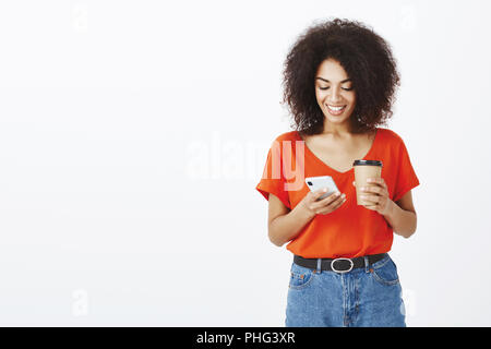 Contrôle de boîte aux lettres en se tenant debout dans la file d'attente. Attrayant moderne femme africaine aux cheveux bouclés en vêtement élégant, holding smartphone et tasse de café, de messagerie tout en regardant l'écran avec sourire Banque D'Images