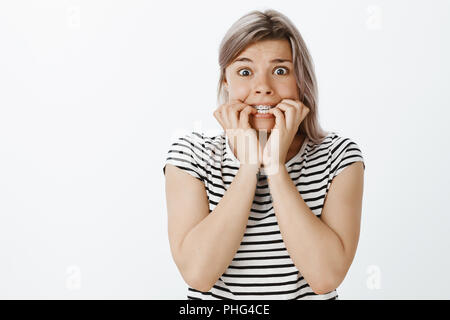 Portrait de peur d'insécurité mignon femme aux cheveux blonds et accolades, mordre les ongles et de peur à l'appareil photo, l'article inquiets et nerveux, peur tout en se posant sur fond gris Banque D'Images