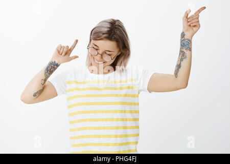 La danse et profiter de la vie. Portrait of happy young positives petite amie en t-shirt à rayures tellow, agitant et faisant des gestes avec les bras tatoués, souriant joyeusement tout en regardant vers le bas, d'être en grande forme Banque D'Images