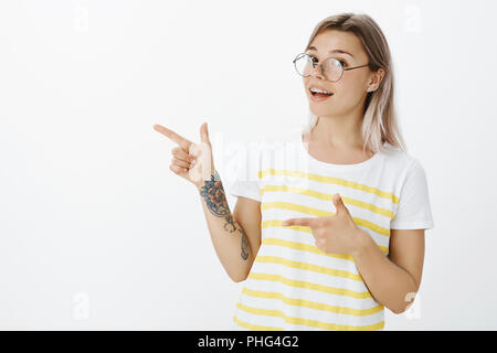 Portrait de smart bon-à femelle en lunettes rondes et les accolades avec bras tatoués, pointant vers la gauche avec des armes à feu et les gestes des doigts tout sourire tout en montrant la direction ou suggérer quelque chose Banque D'Images