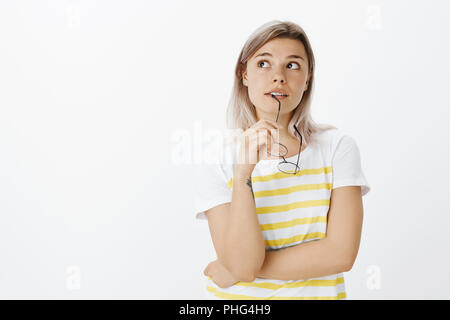 Tourné à l'intérieur de rêve bon-à la femme blonde en tenue décontractée, mordre et lunettes de rim à la recherche dans le coin en haut à gauche en pensant, faire des choix ou de douter de l'esprit, posant sur mur gris Banque D'Images