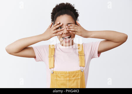 Girl peeking avec un œil à don père prépare pour elle. Portrait de femme charmante et enfantine sans soucis avec la peau foncée et les cheveux bouclés, couvrant les yeux avec les paumes et souriant joyeusement Banque D'Images