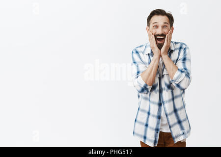 Étant père de famille impressionné lui a rendu visite sur b-jour. Portrait de surprise ravie et amusée homme debout sur fond gris, heureux et joyeux, ne peut pas croire ses yeux, tenant paumes sur les joues Banque D'Images