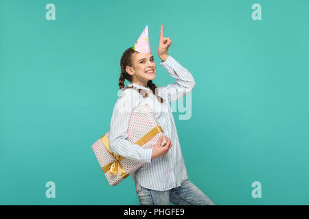 Happy Funny Girl en chemise bleue à rayures avec un permanent et boîte-cadeau anniversaire cap sur sa tête, looking at camera avec safisfied smiley face, Indoo Banque D'Images