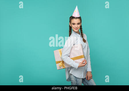 Happy Funny Girl en chemise bleue à rayures avec un permanent et boîte-cadeau anniversaire cap sur sa tête, looking at camera avec safisfied smiley face, Indoo Banque D'Images