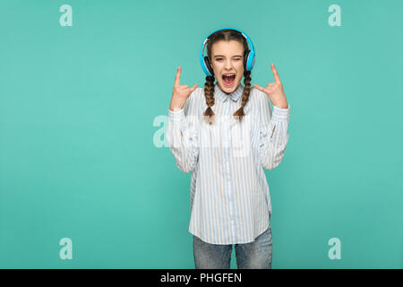 Heureux surpris fille en chemise bleu rayé d'écoute permanent musique préférée avec casque, looking at camera avec visage surpris et rock signe, Piscine Intérieure Banque D'Images