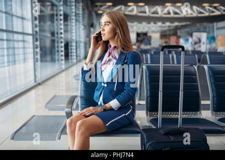 Hôtesse de l'air avec valise assis sur son fauteuil dans la salle d'attente dans l'aéroport et de parler par téléphone mobile. Hôtesse de l'air, avec des bagages, avec la main de bord Banque D'Images