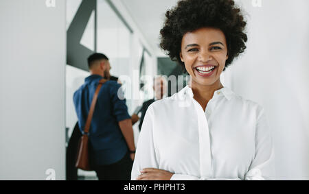 Portrait of happy young African businesswoman looking at camera et souriant avec son équipe à discuter en arrière-plan. Cheerful woman in office hallway w Banque D'Images