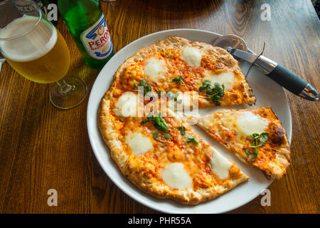 Pizza Margherita classique de fromage mozzarella sur une plaque blanche avec une tranche découper servi avec une bouteille et un verre de la bière italienne Peroni Banque D'Images