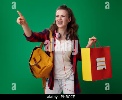 Souriante jeune femme étudiant dans un chandail rouge avec sac à dos et casque holding Shopping bag avec des livres et du doigt quelque chose sur fond vert Banque D'Images