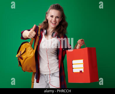 Heureux jeune étudiante femme dans une chemise rouge avec sac à dos et casque holding Shopping bag avec livres et showing Thumbs up sur fond vert Banque D'Images